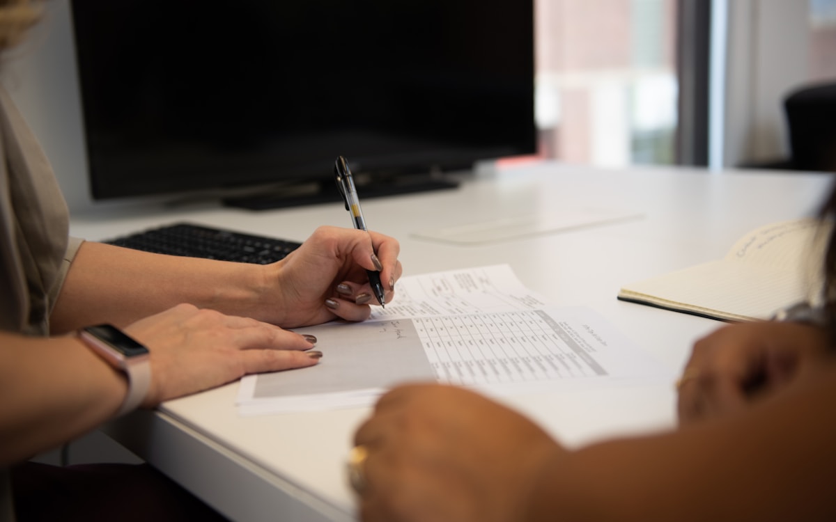 Zwei Hände am Schreibtisch füllen gemeinsam ein Formular mit Stift aus, helles Büro.
