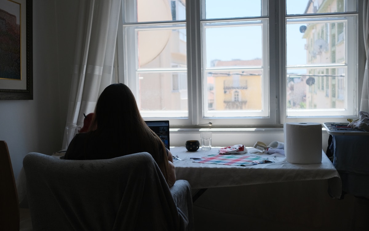 Frau arbeitet am Laptop an einem hellen Fenster im Wohnraum, Tageslicht.