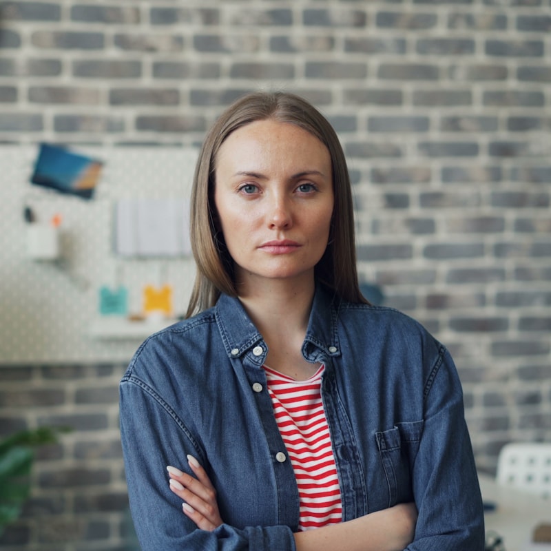 Symbolbild: Frau in heller Bluse mit verschränkten Armen vor Bürohintergrund. Echtes Foto folgt.